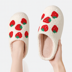 Pair of cream-colored slippers with red strawberry pattern held by a hand on a white background